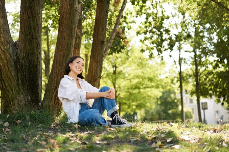 Beautiful smiling girl sits near tree in park, enjoying nature outdoors, relaxing and resting on fresh air. Copy space