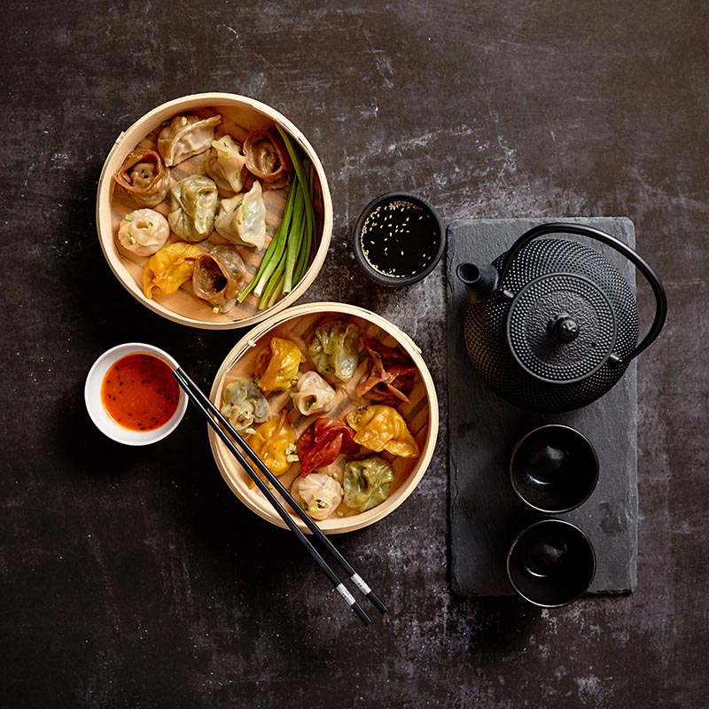 Composition of chinese food. Mixed kinds of dumplings from wooden bamboo steamer. Served with soy and spicy sauces and traditional tea in iron pot. Top view with copy space.