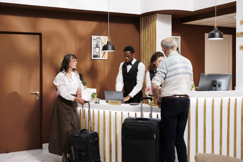 Male and female elderly guests in modern hotel lobby, with attentive staff providing efficient assistance during check-in and reservation procedures. Senior tourists wait with suitcases at front desk.